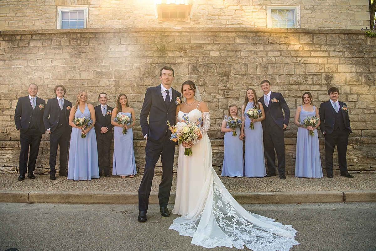 Grace, Alex, and their wedding party pose along a historic stone wall in downtown Ste. Genevieve