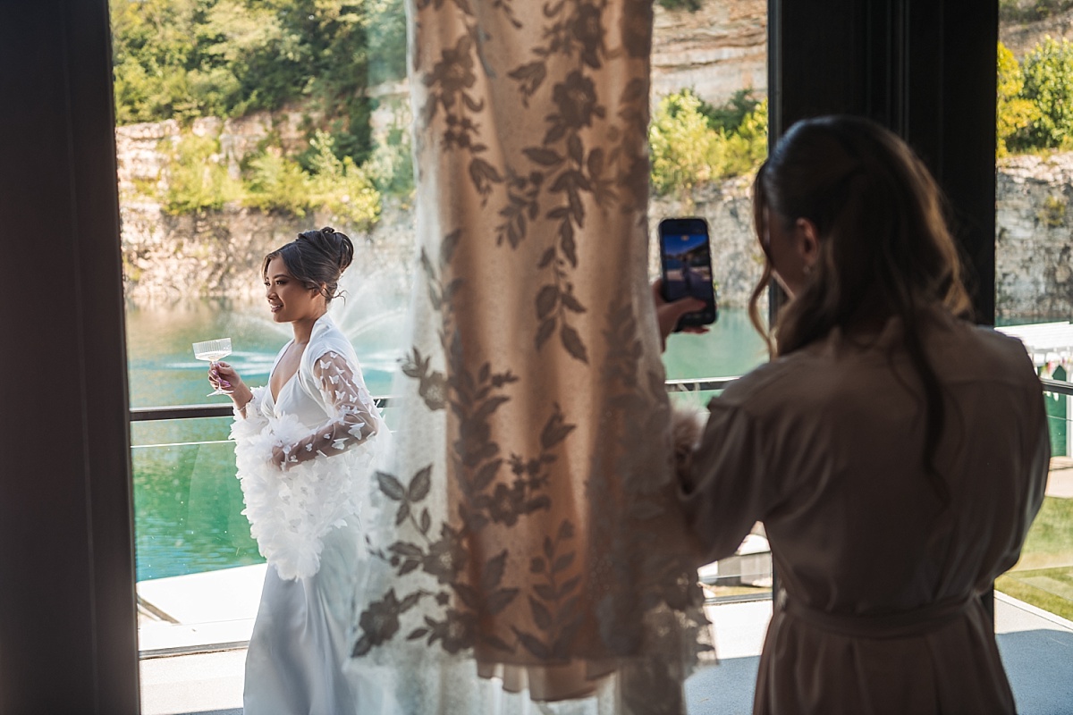 Melicia holds a champagne glass by the lake while a friend photographs her through the window.