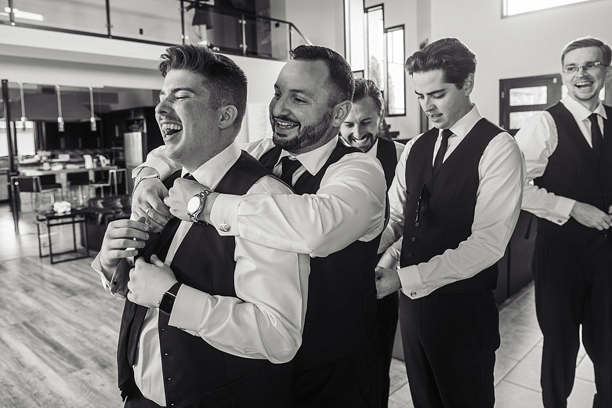 Justin laughs with his groomsmen as they help him get ready in a candid black-and-white photograph.