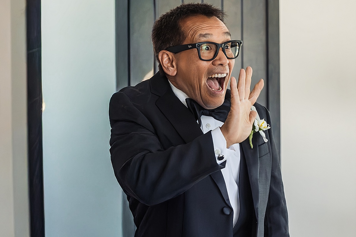 A wedding guest in a black tuxedo and glasses reacts with joyful excitement.