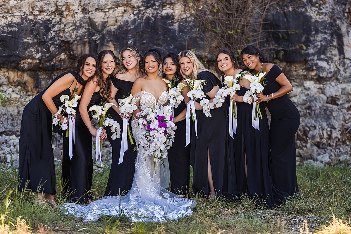 Melicia smiles with her bridesmaids in black dresses, holding white and purple orchid bouquets.