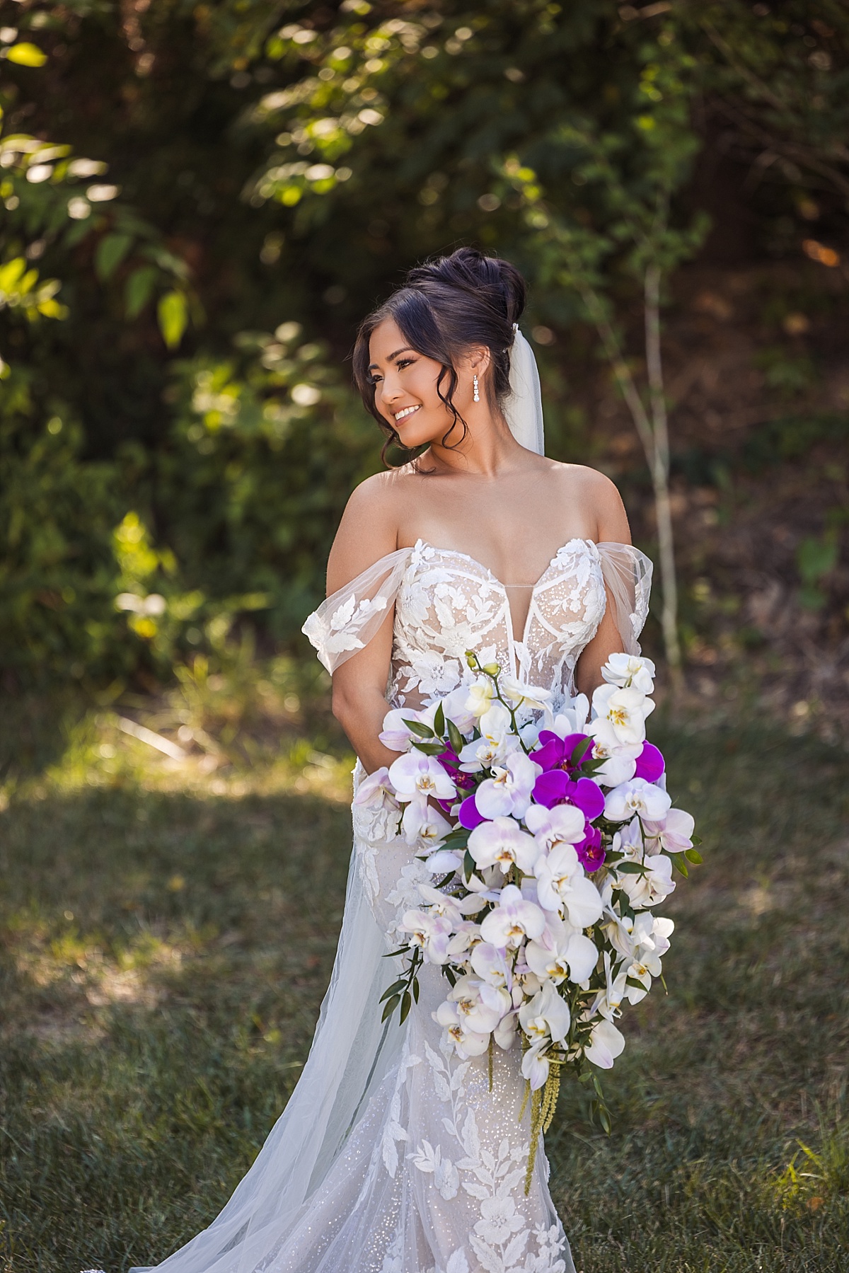 Melicia smiles in her lace wedding gown while holding a cascading white and purple orchid bouquet.