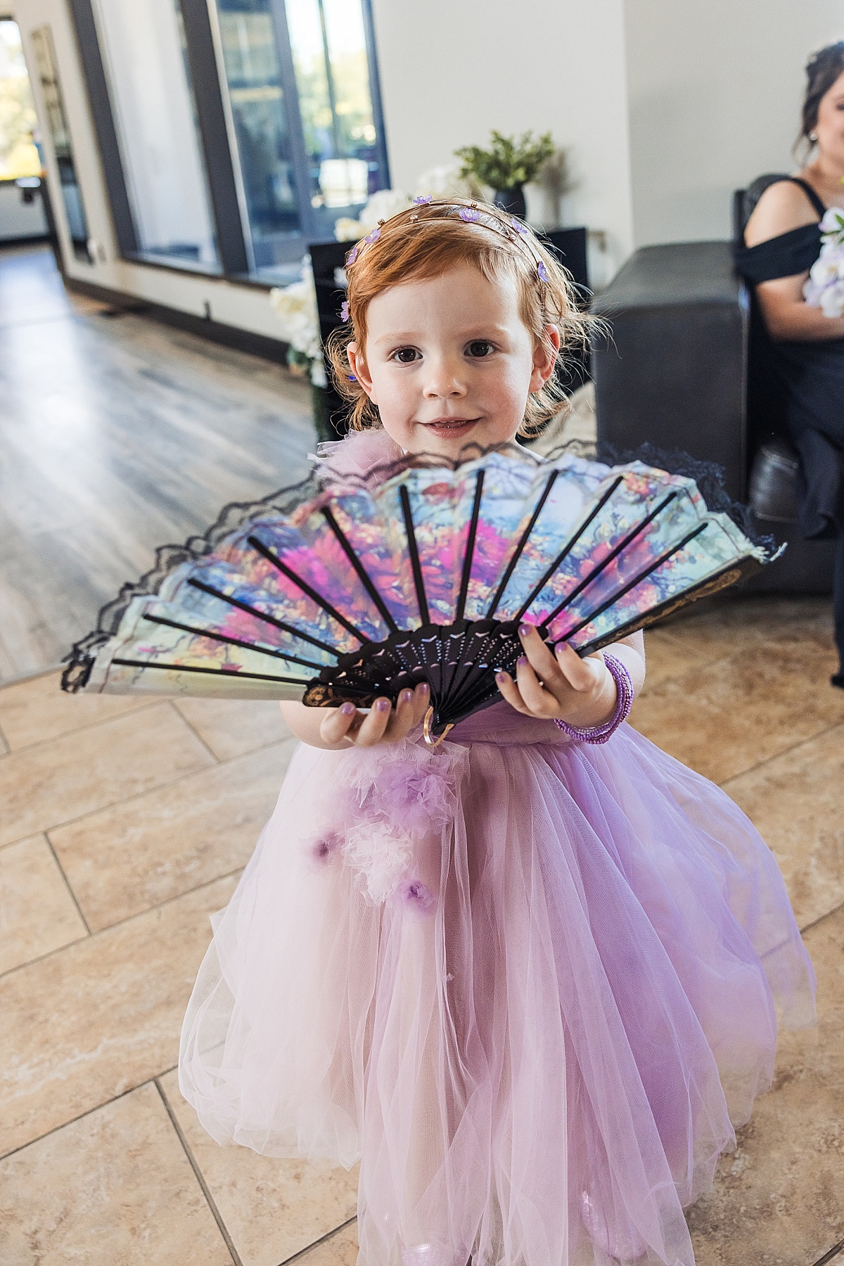 A young flower girl in a lavender dress holds an open decorative fan at the wedding reception.
