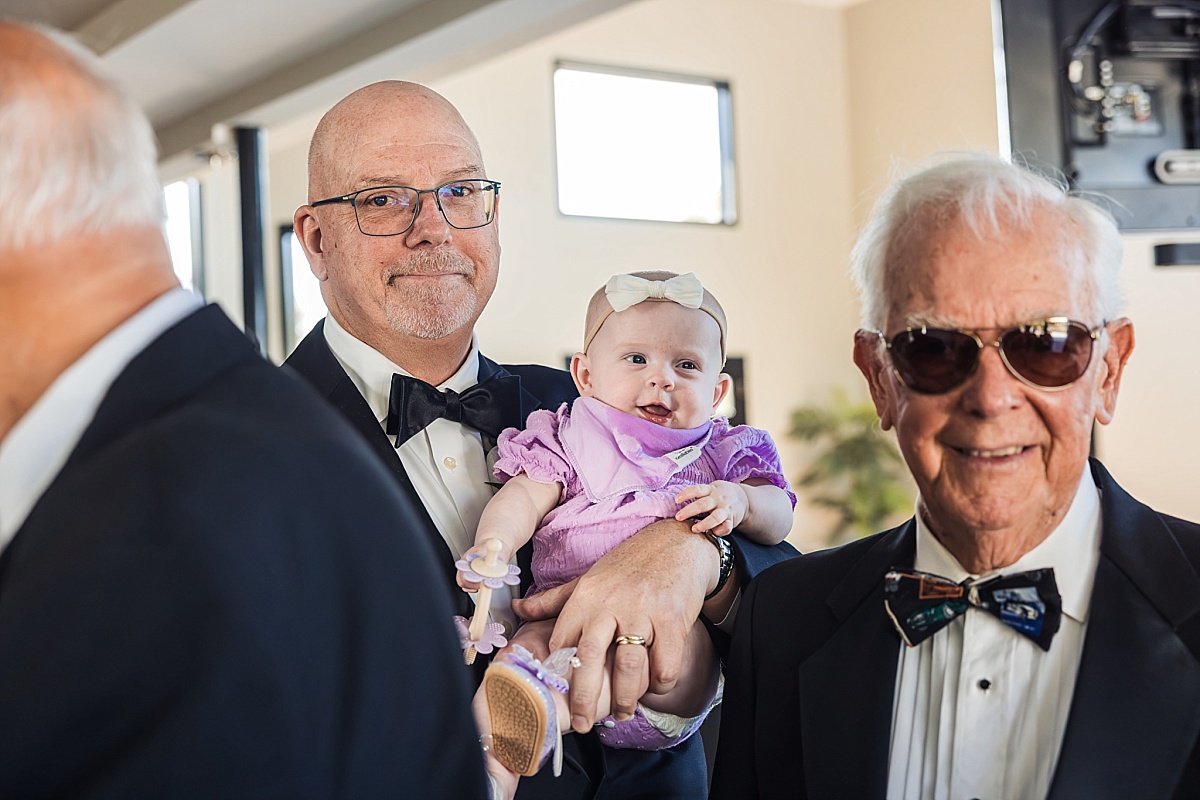 Three generations of family members share a moment together, with a baby in a lavender outfit held by a wedding guest.