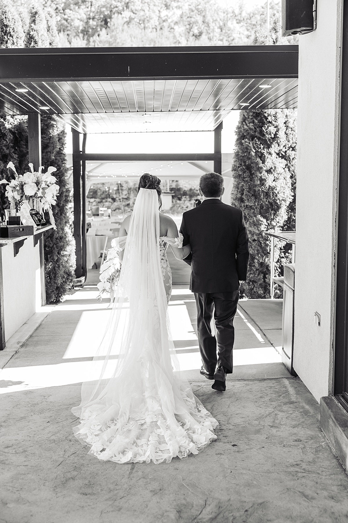 Black-and-white photograph of Melicia walking toward the ceremony with her father, her long veil and lace train flowing behind her.