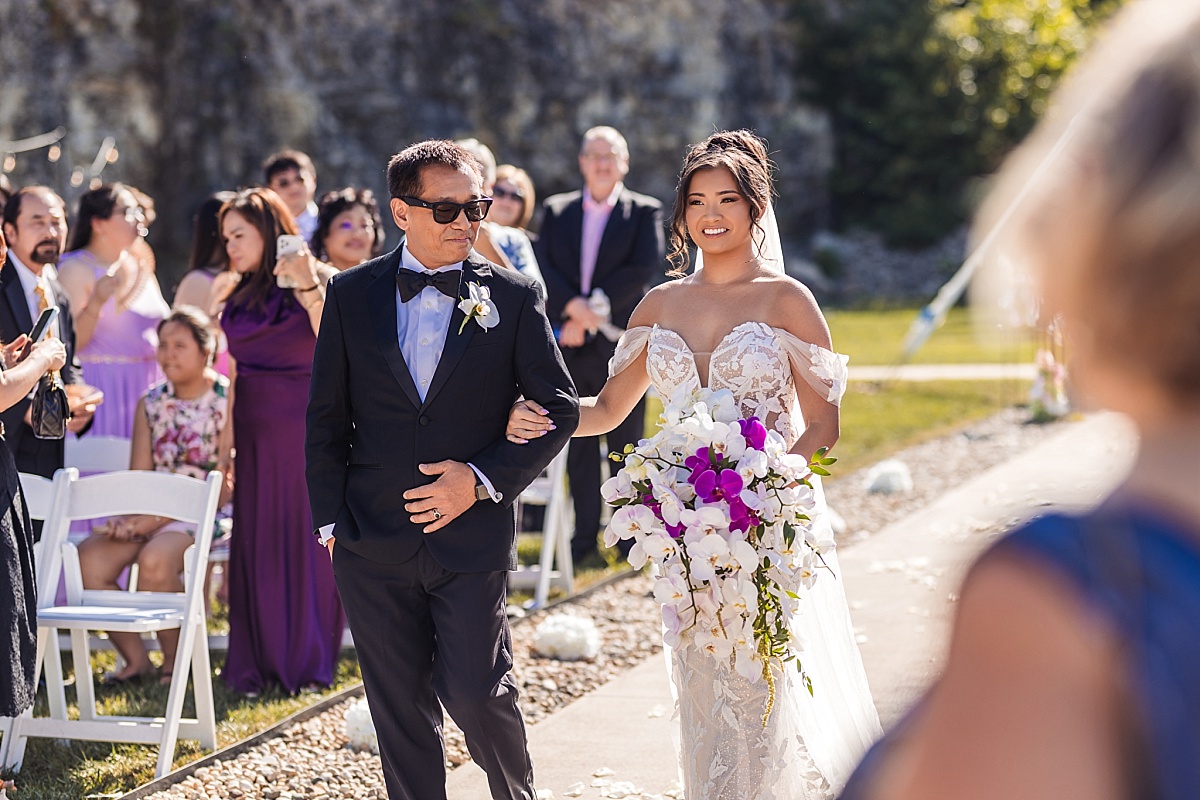 Melicia walks arm-in-arm with her father toward the outdoor wedding ceremony as guests look on.