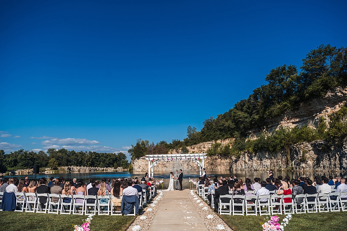 Wide view of the outdoor wedding ceremony at Sunset Bluffs, with guests seated beside the lake and limestone cliffs.
