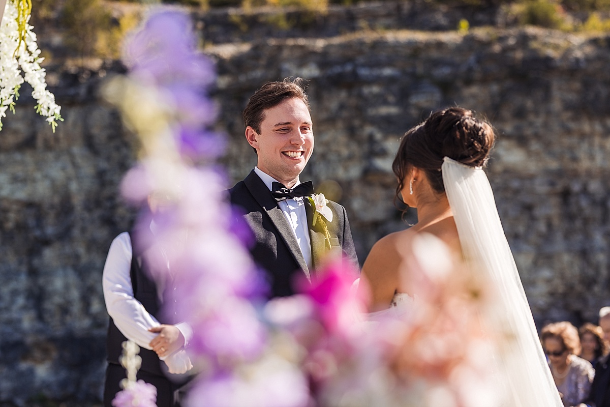 Justin smiles at Melicia during their outdoor wedding ceremony, with purple flowers softly framing the foreground.