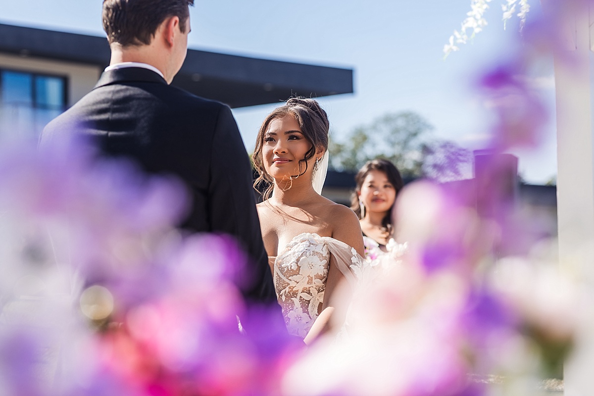 Melicia looks lovingly at Justin during their outdoor wedding ceremony, framed by soft purple flowers.