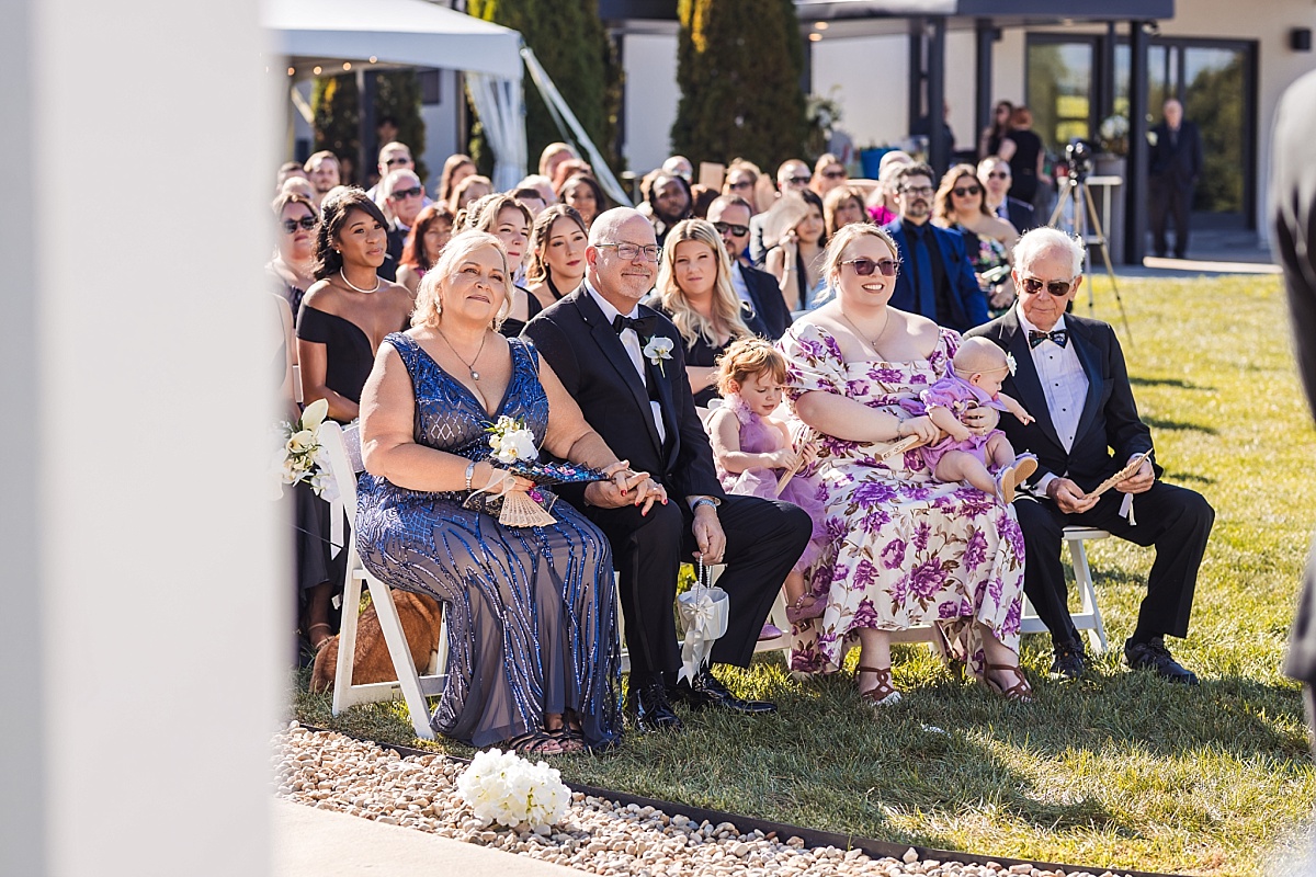Family and friends watch Melicia and Justin's wedding ceremony from rows of white chairs beside the lake.