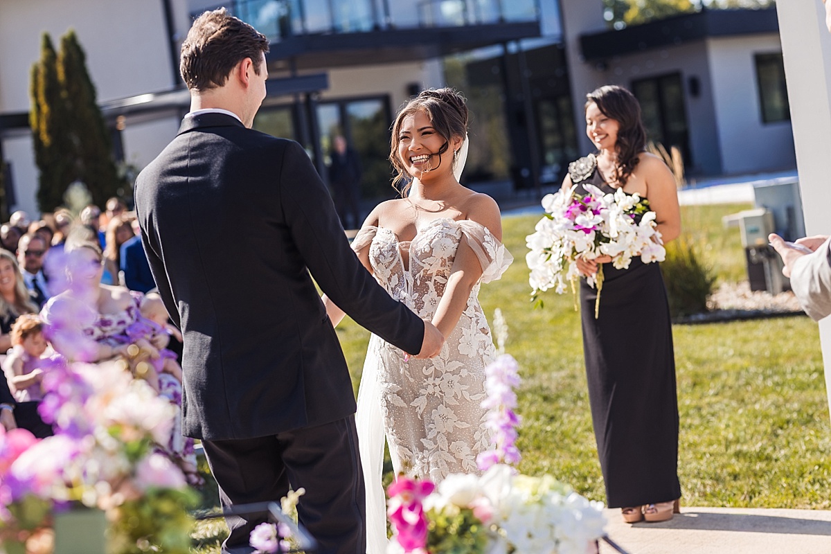 Melicia beams at Justin during the ceremony as her bridesmaid stands nearby with a bouquet.