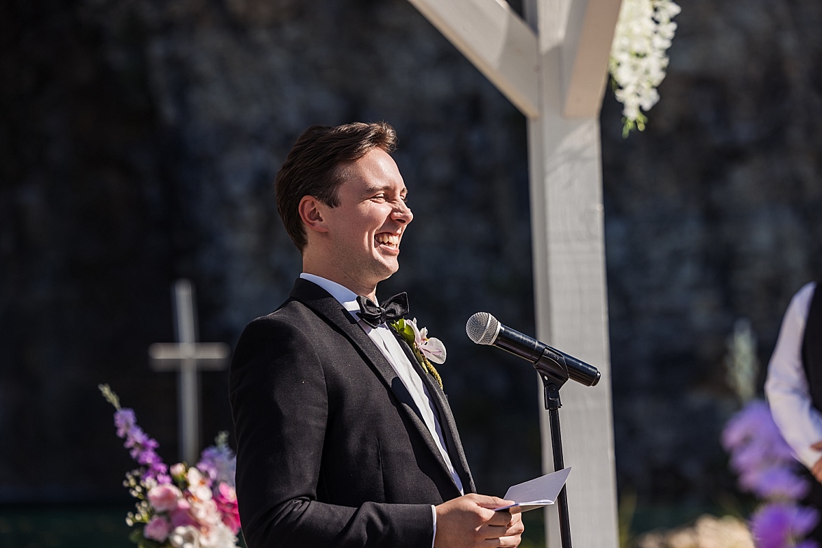 Justin laughs during his wedding ceremony, holding his vows beside the flower-decorated ceremony structure.