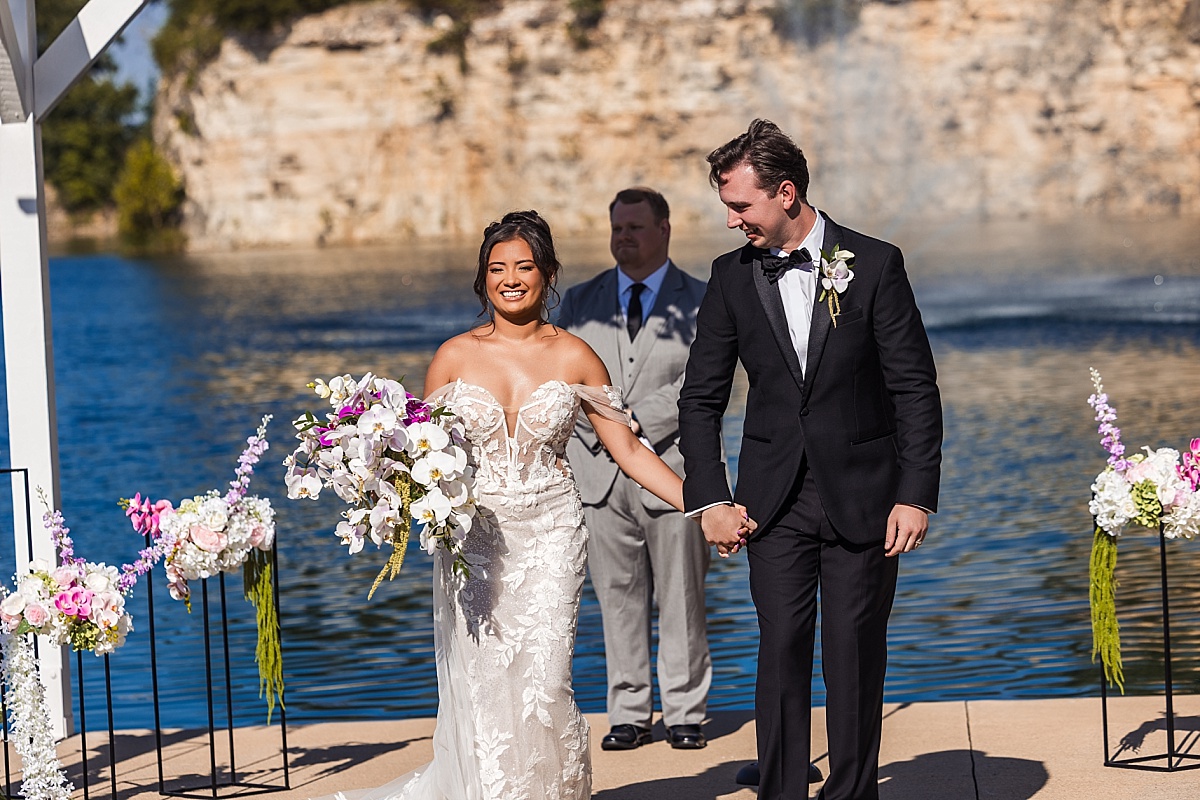 Melicia and Justin walk hand-in-hand away from their outdoor ceremony, smiling in front of the lake and stone bluffs.