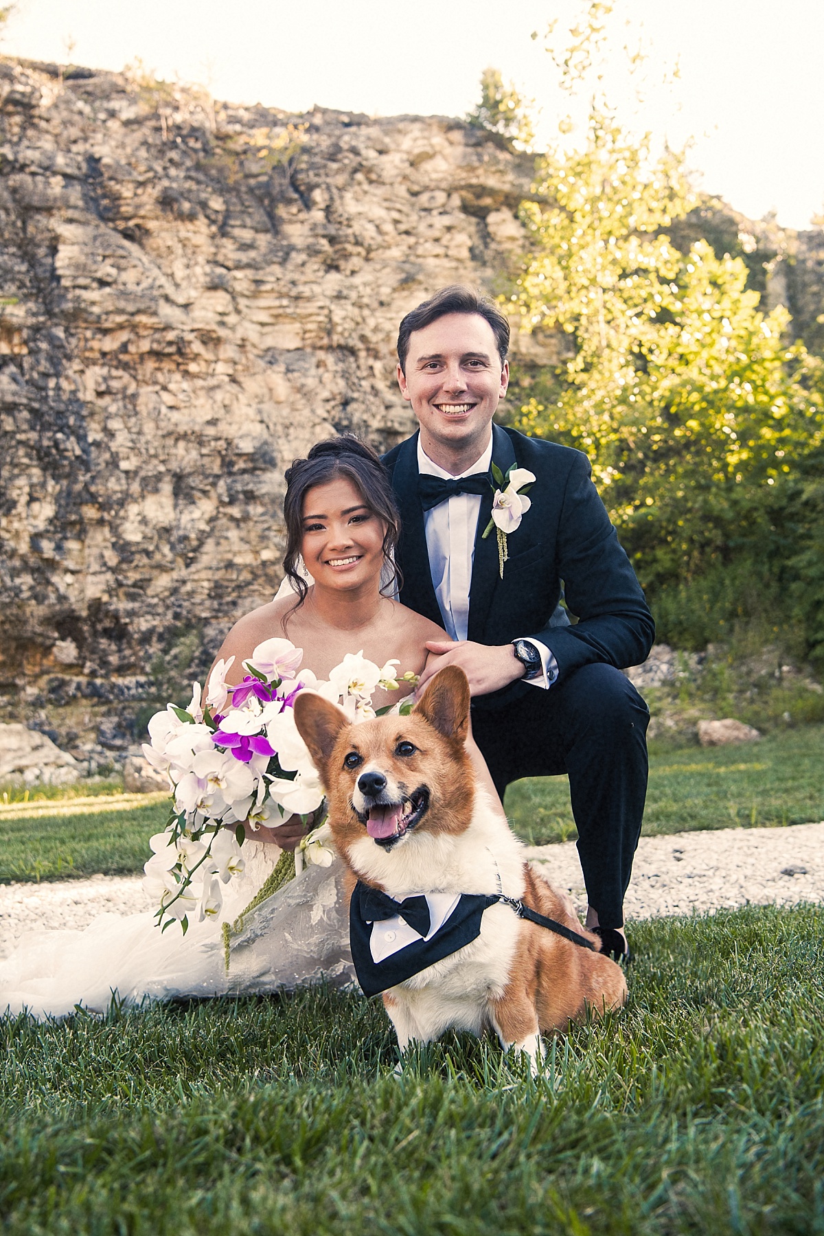 Melicia and Justin pose with their corgi in front of the stone bluffs, with the dog wearing a tiny black tuxedo.