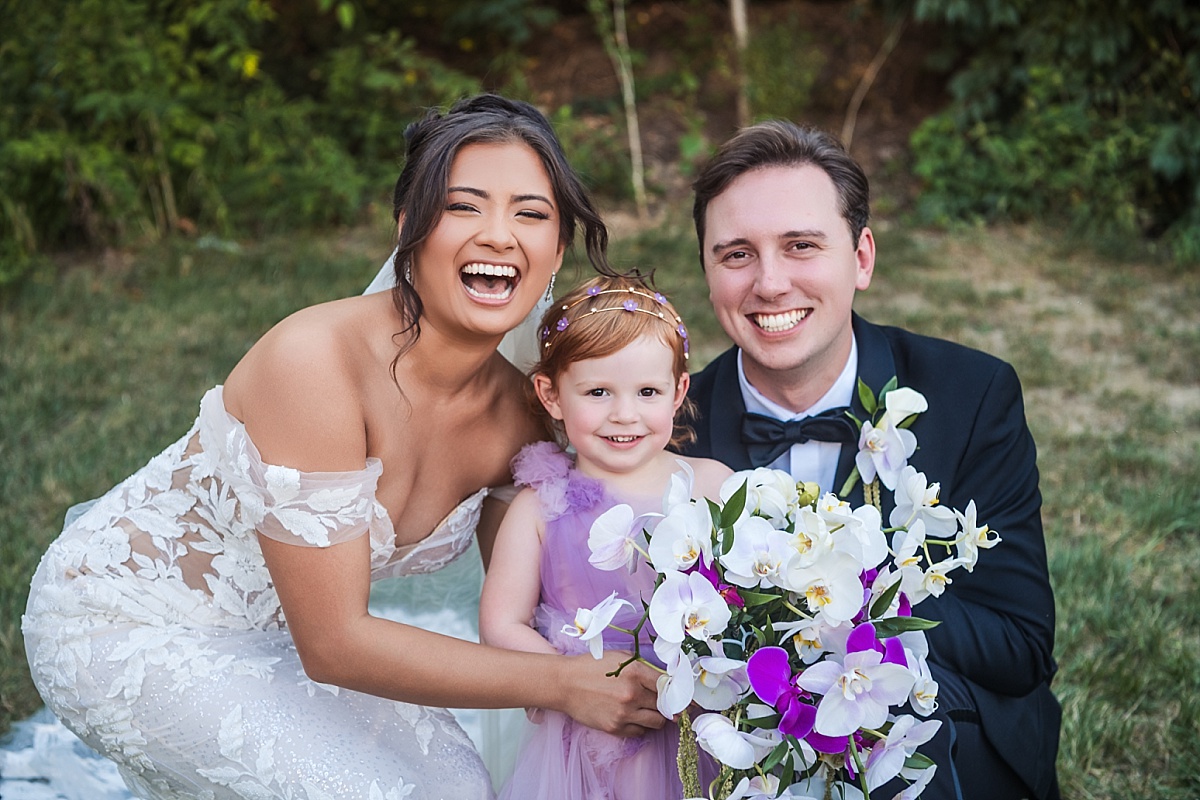 Melicia and Justin crouch beside a young flower girl in a lavender dress for a joyful family portrait.