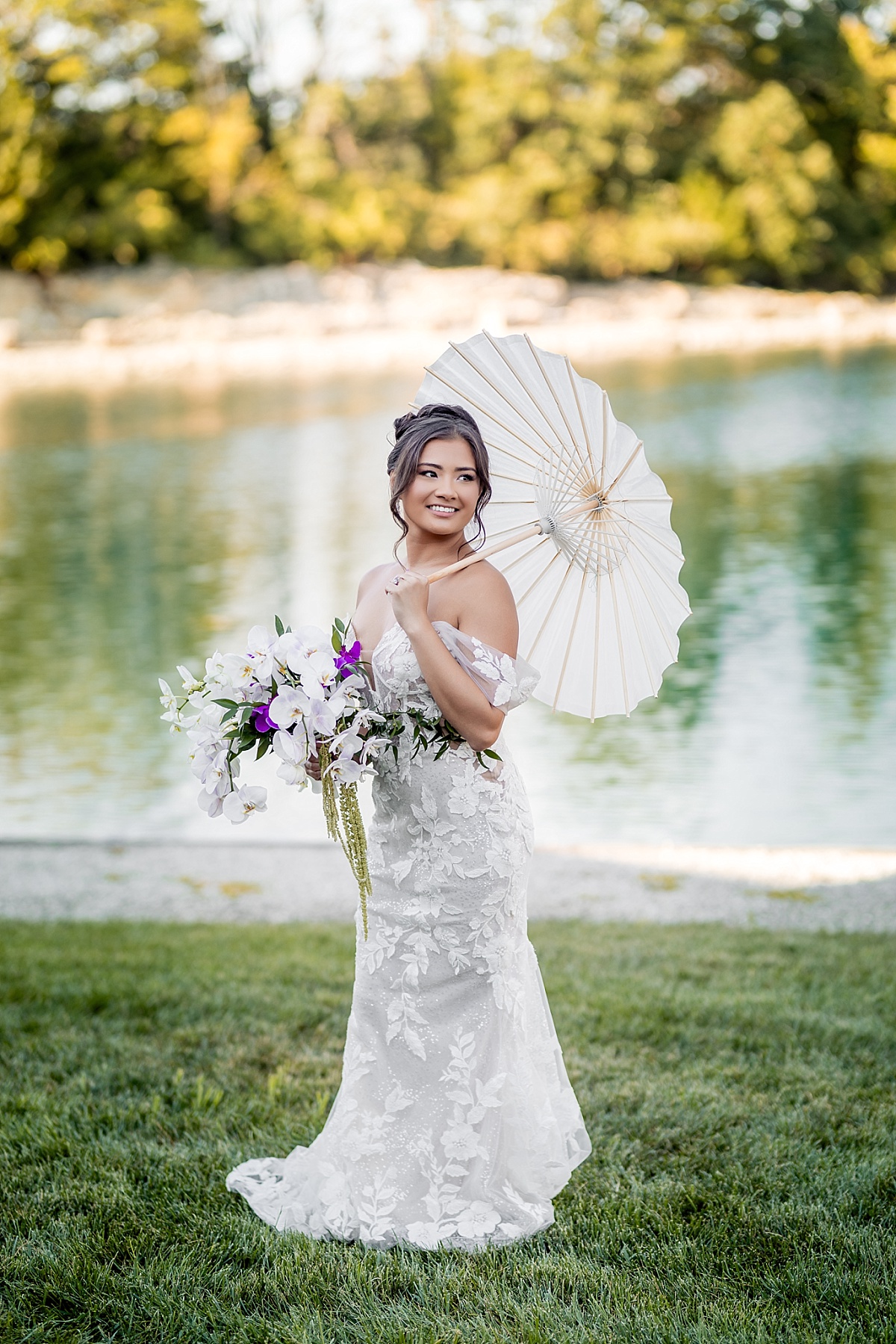 Melicia poses beside the lake in her lace wedding gown, holding her orchid bouquet and a white parasol.