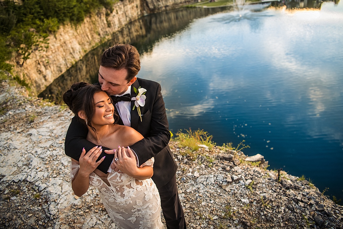Justin kisses Melicia's forehead as they embrace beside the lake in the golden evening light.
