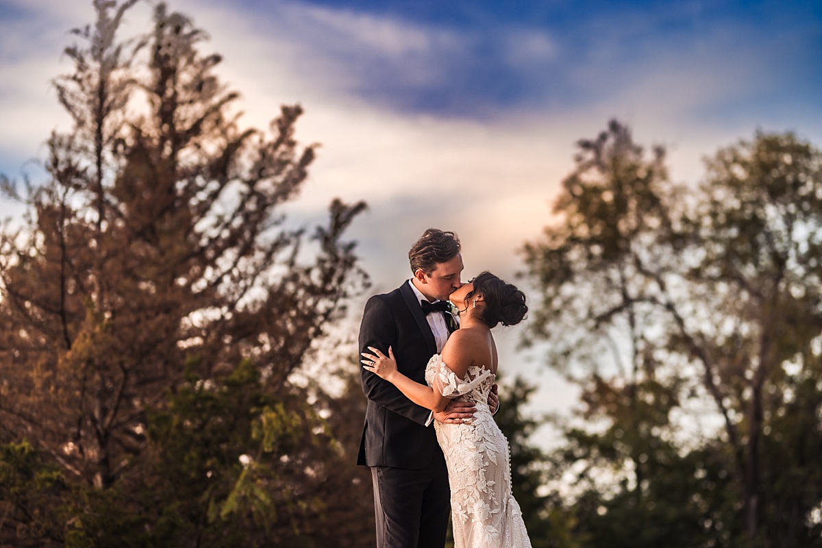 Melicia and Justin kiss at sunset, silhouetted against the colorful sky and trees.