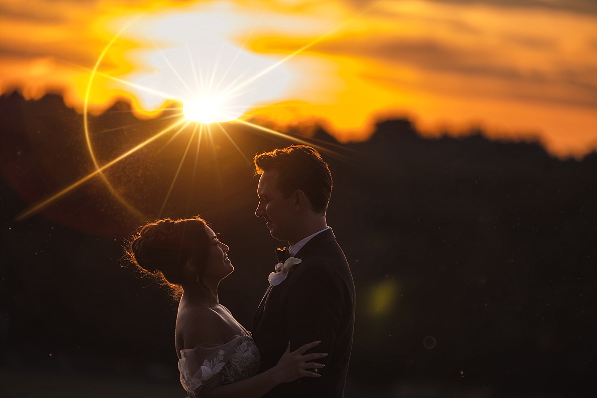Melicia and Justin stand close together in silhouette beneath a vivid orange sunset.