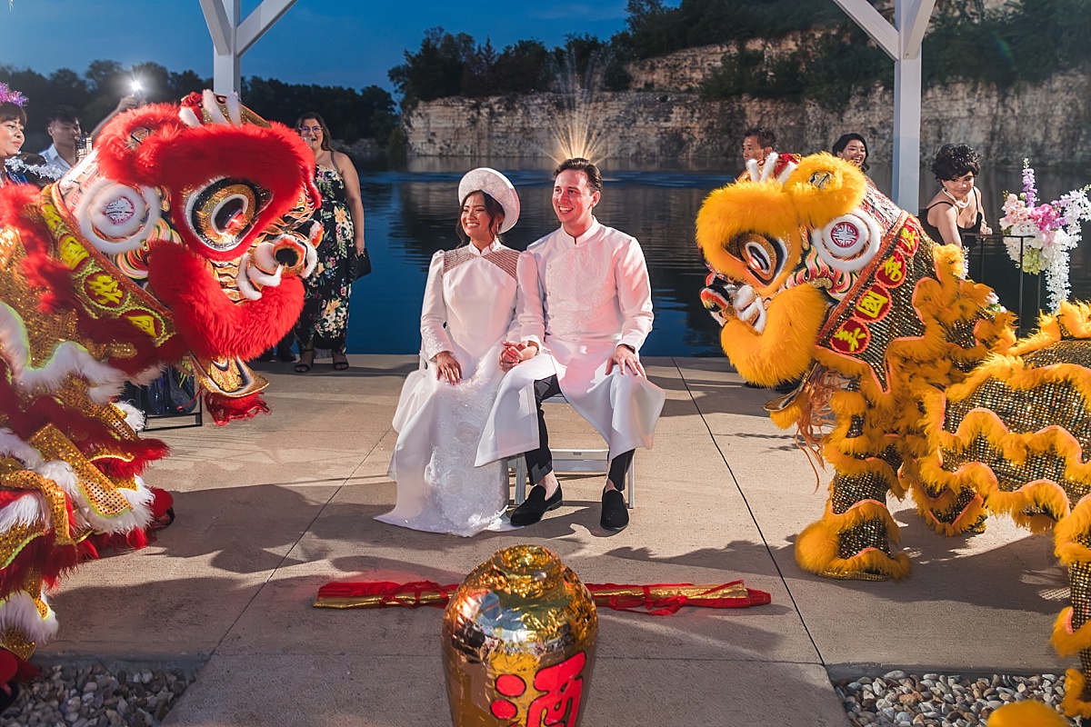Melicia and Justin sit together in traditional Vietnamese wedding attire as red and yellow lion dancers perform beside the lake.
